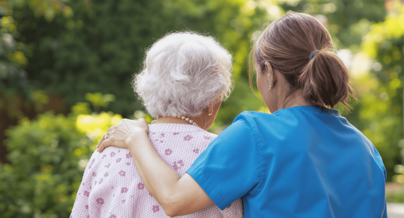 Caregiver with her arm around an elderly woman in a garden.