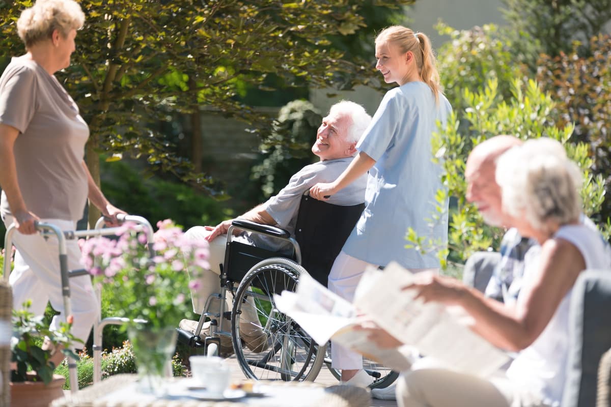 Caregiver pushing an elderly man in a wheelchair in a sunny garden, with other residents nearby.